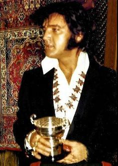 1970 Elvis in black jacket holding silver chalice cup rug hung in background